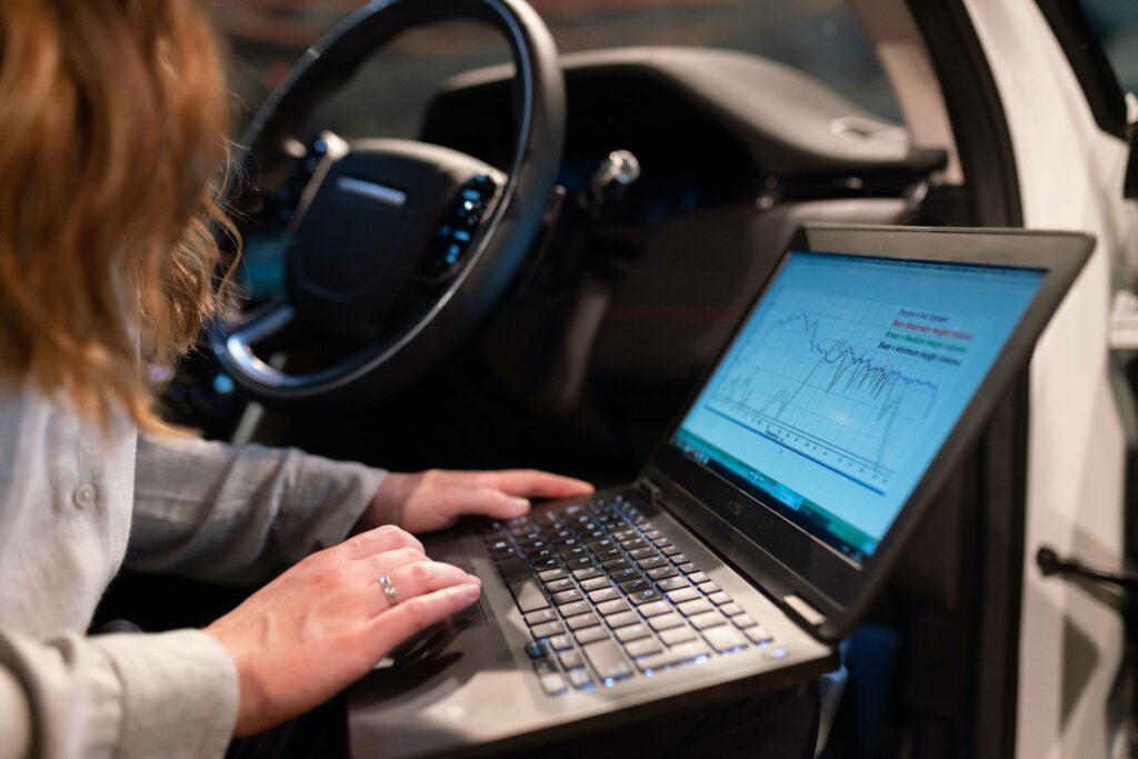 Frau arbeitet an einem Laptop im Auto, auf dem Bildschirm werden Fahrzeugdiagnosedaten und Diagramme angezeigt.